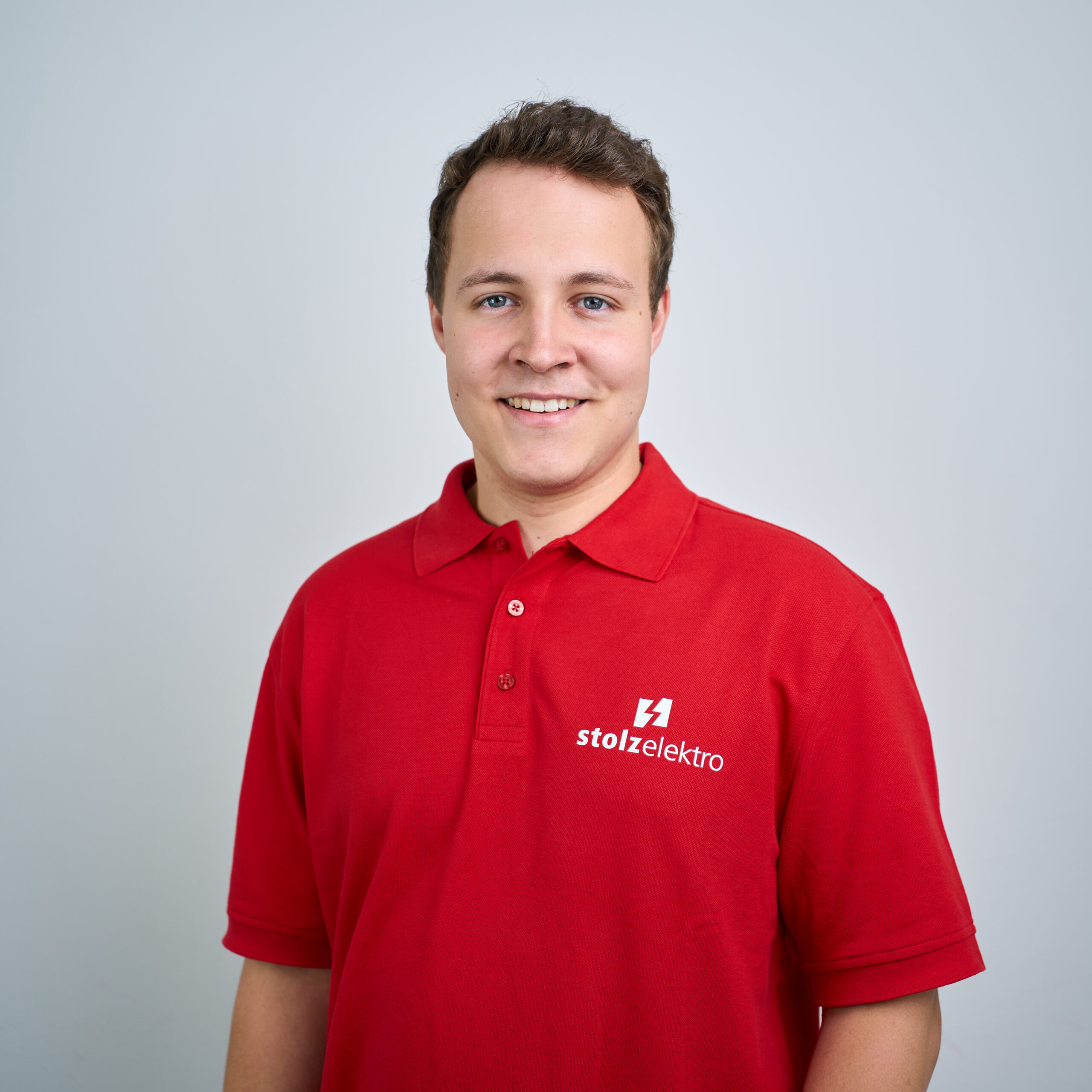Eine lächelnde Person, die ein rotes Poloshirt mit dem stolzelektro-Logo trägt, steht vor einem schlichten hellgrauen Hintergrund.