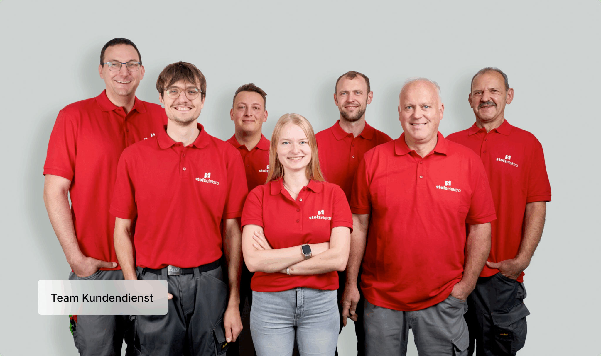 Sieben Personen in roten Poloshirts mit einem Firmenlogo stehen lächelnd vor einem hellgrauen Hintergrund. Eine Aufschrift auf dem Bild lautet "Team Kundendienst". Die Gruppe besteht aus zwei Männern und einer Frau.
